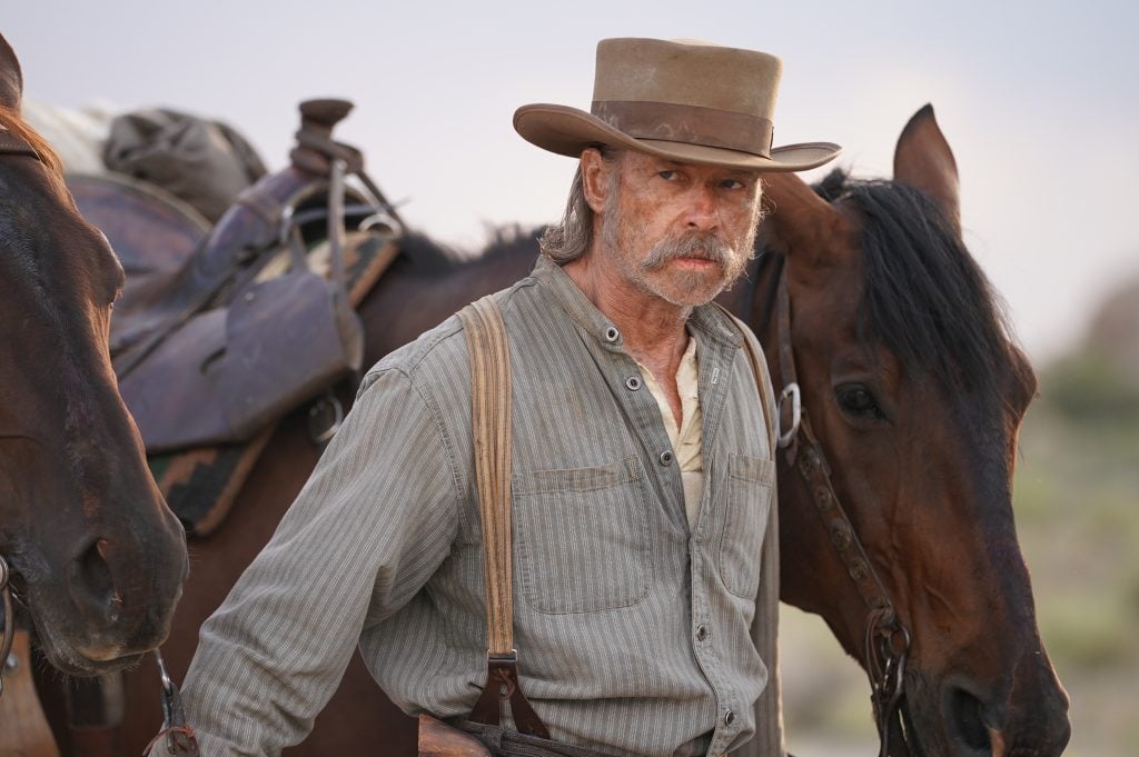 A man in vintage Western attire with suspenders and a wide-brimmed hat stands between two horses outdoors.
