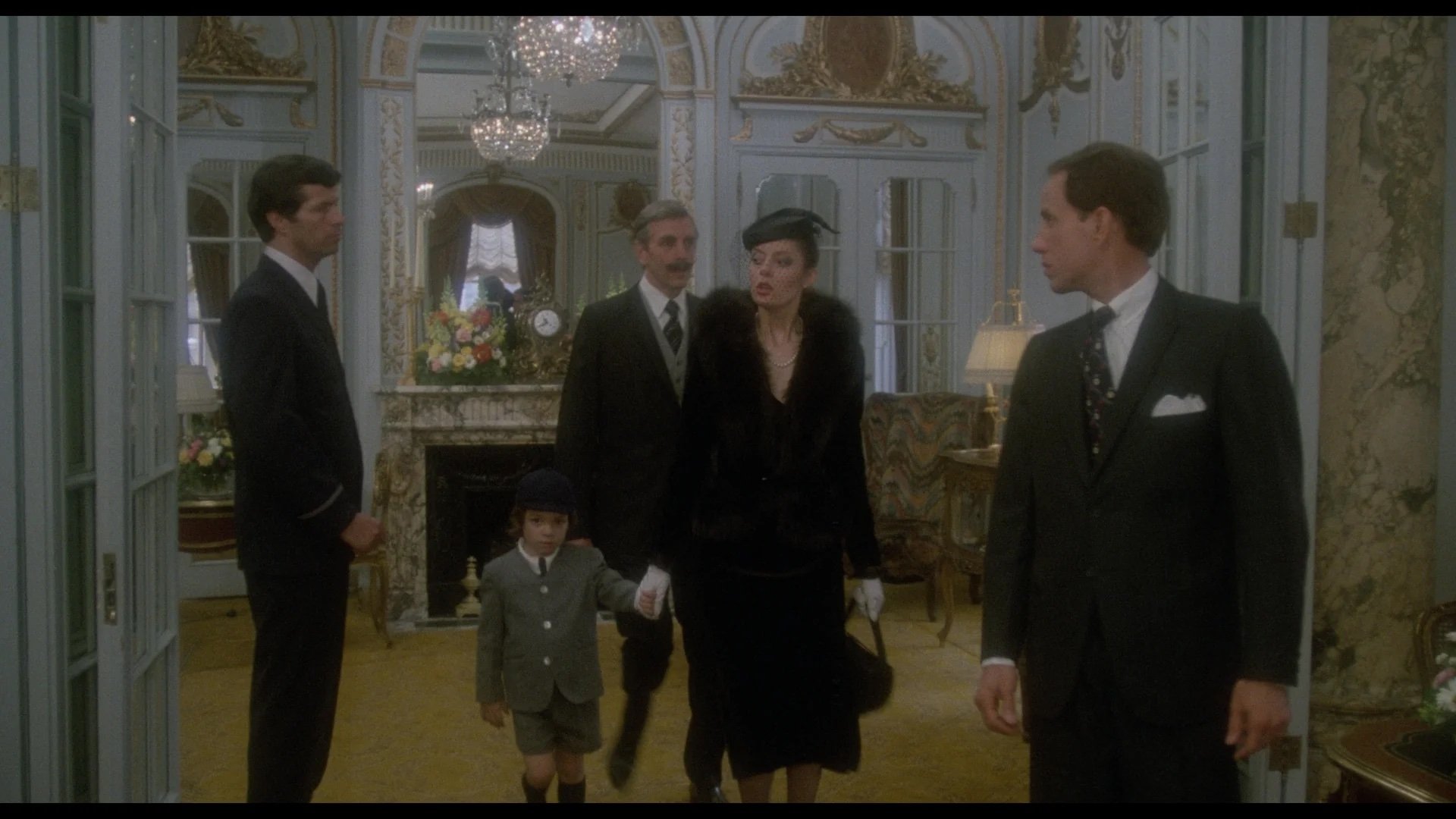 Five people, including a child, stand in a formal, ornately decorated room with chandeliers, mirrors, and a fireplace. The adults wear formal clothing, and the child is dressed in a suit and hat.