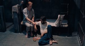 Two men sit on the floor and bed in a dimly lit prison cell, with snacks and playing cards between them, one reaching out to the other.