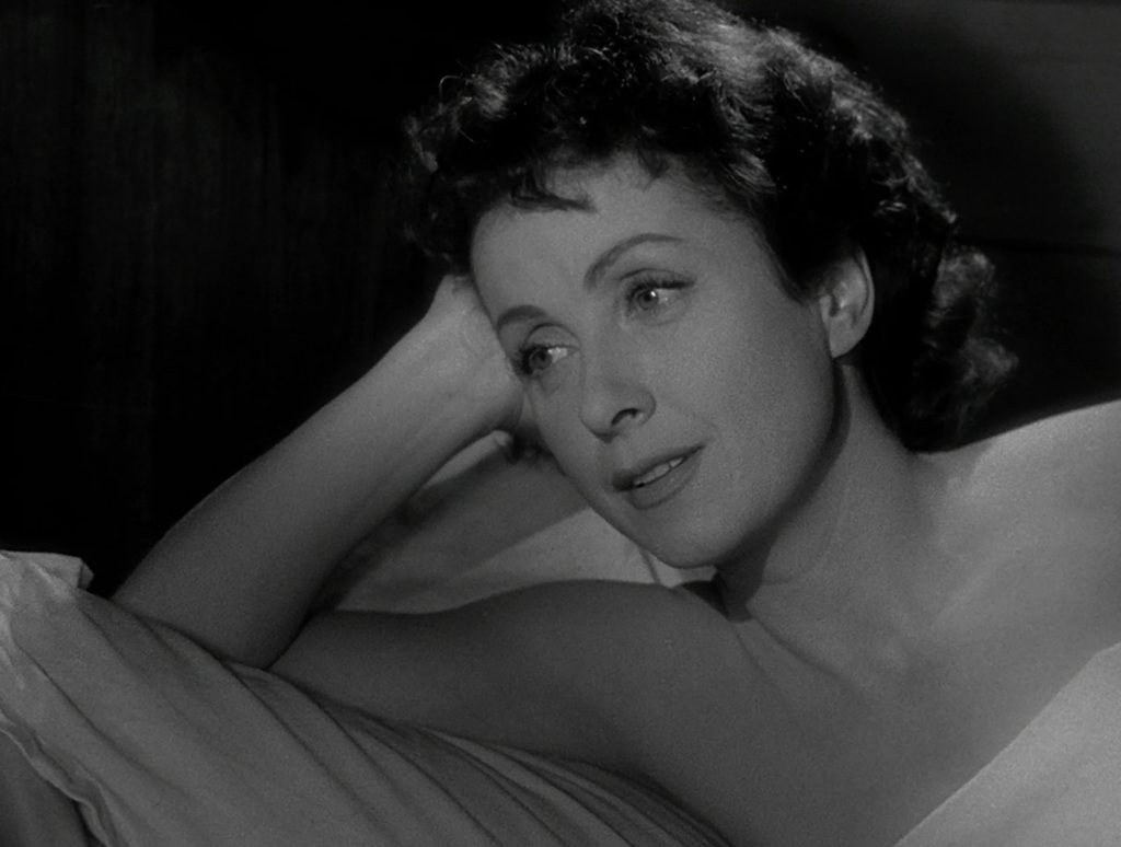 A woman with short, curly dark hair lies on a pillow, propping her head up with her hand, and gazes thoughtfully to the side in a black and white image.