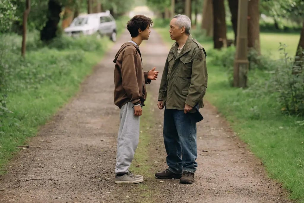 Two people stand facing each other on a tree-lined path, engaged in conversation; a parked car is visible in the background.