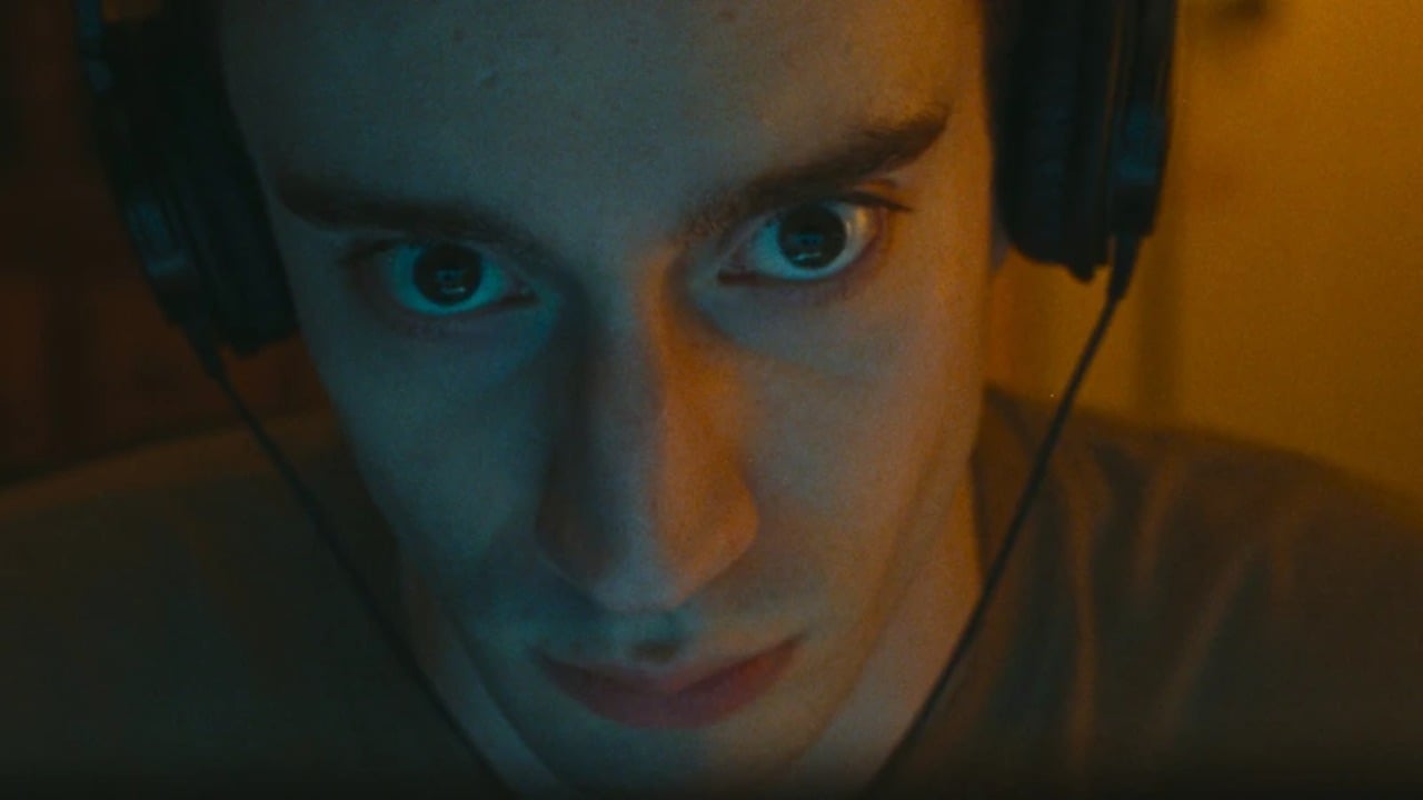 A person wearing headphones looks directly at the camera with a serious expression in low, warm lighting.