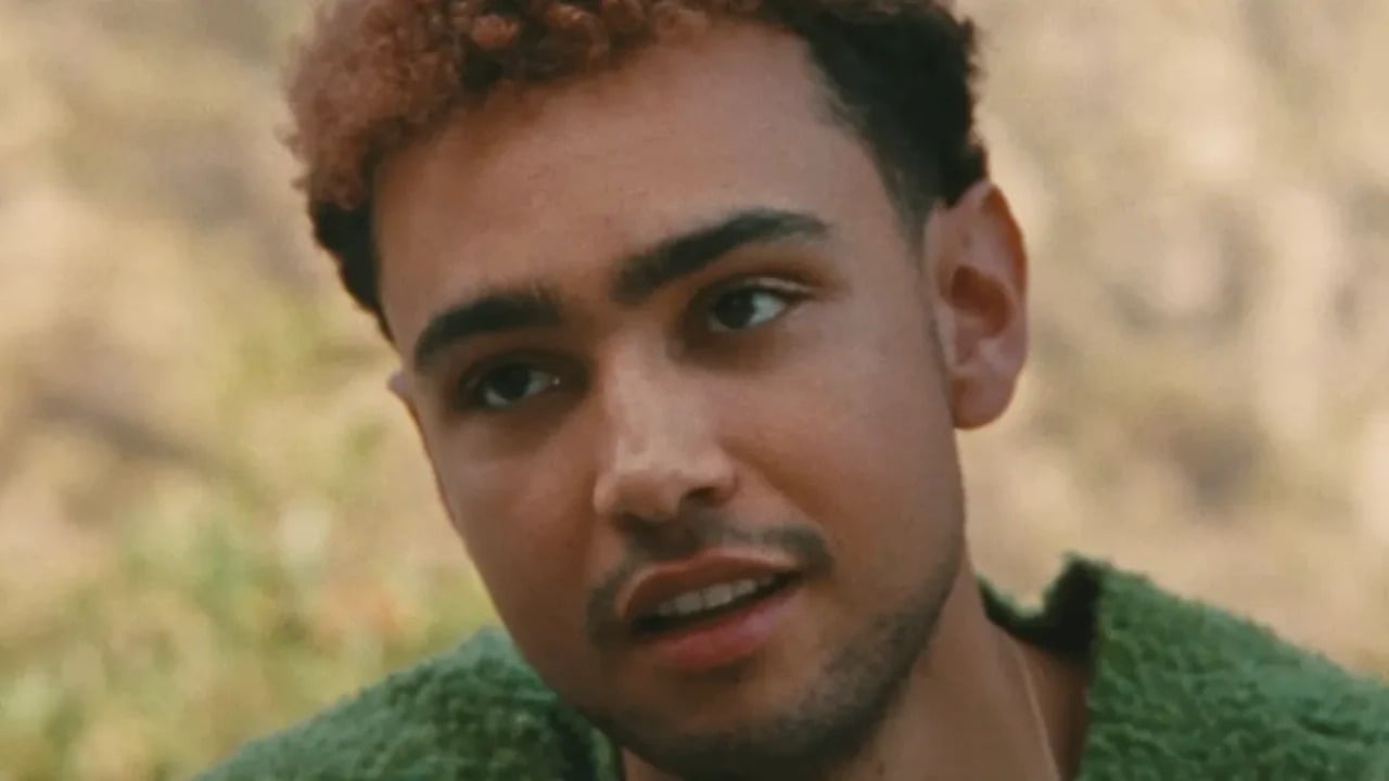 A young person with short, curly brown hair and light facial hair looks slightly to the side, wearing a green textured sweater outdoors.