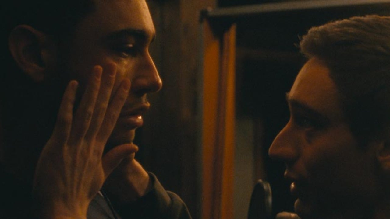 Two men face each other in dim lighting; one touches the other's face gently, their expressions serious and focused.