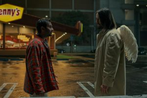 Two men stand face-to-face in a parking lot at night; one wears a plaid shirt, the other a trench coat with angel wings. A Denny’s restaurant sign is visible in the background.