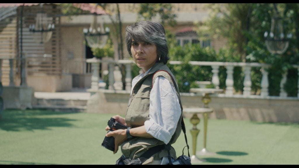 A person in a vest holds a camera and looks toward the viewer while standing outdoors on a lawn, with trees and a building in the background.
