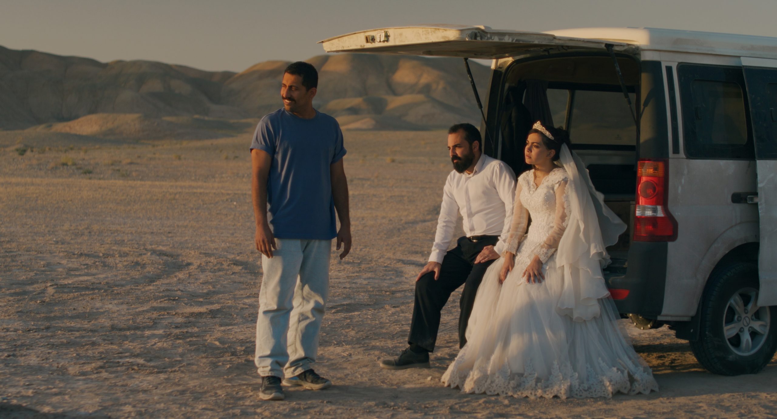 A bride and groom sit at the open back of a vehicle in a desert landscape while a man in casual clothes stands nearby. Mountains are visible in the background.