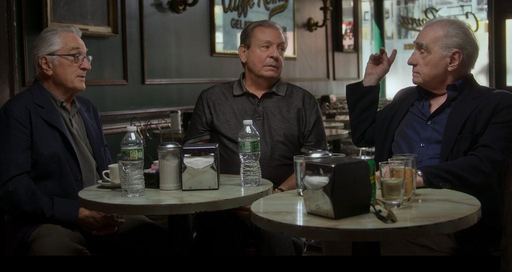 Three older men sit around small café tables having a conversation, with water bottles and cups on the tables in front of them.