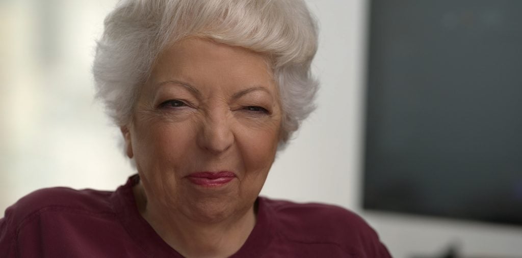 Older woman with short white hair wearing a maroon top, squinting and smiling slightly, in an indoor setting.