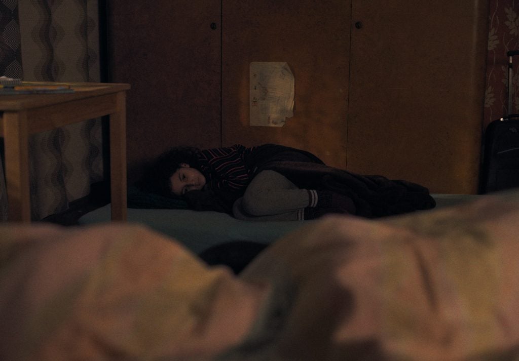 A person lies curled up on a makeshift bed on the floor, partially covered by a blanket, beside a wooden table in a dimly lit room.