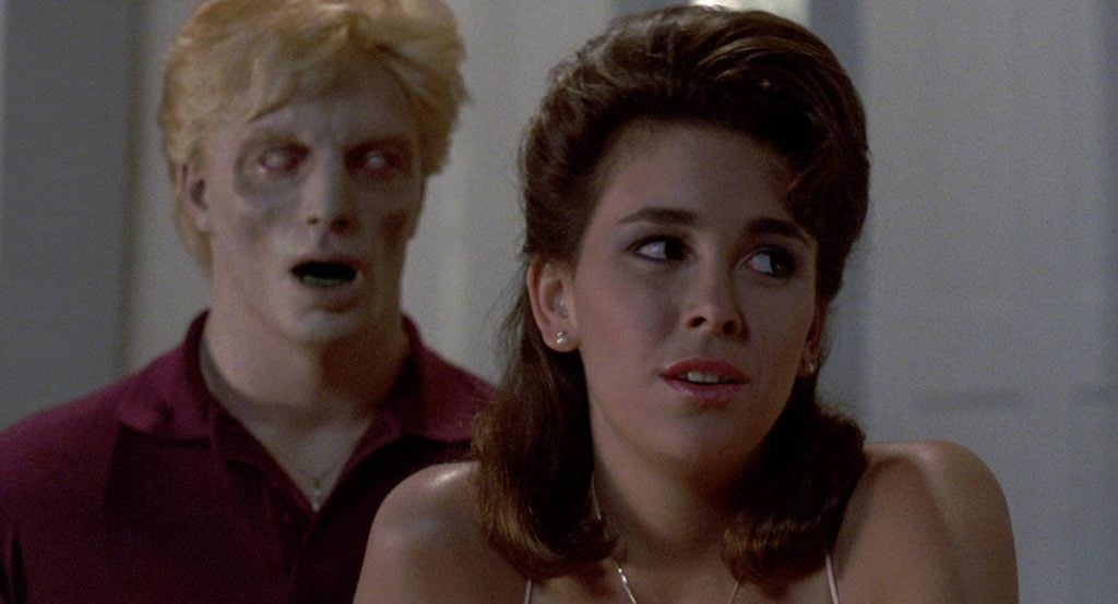 A woman looks concerned while a man with pale skin and zombie-like features stands behind her, mouth slightly open, indoors.