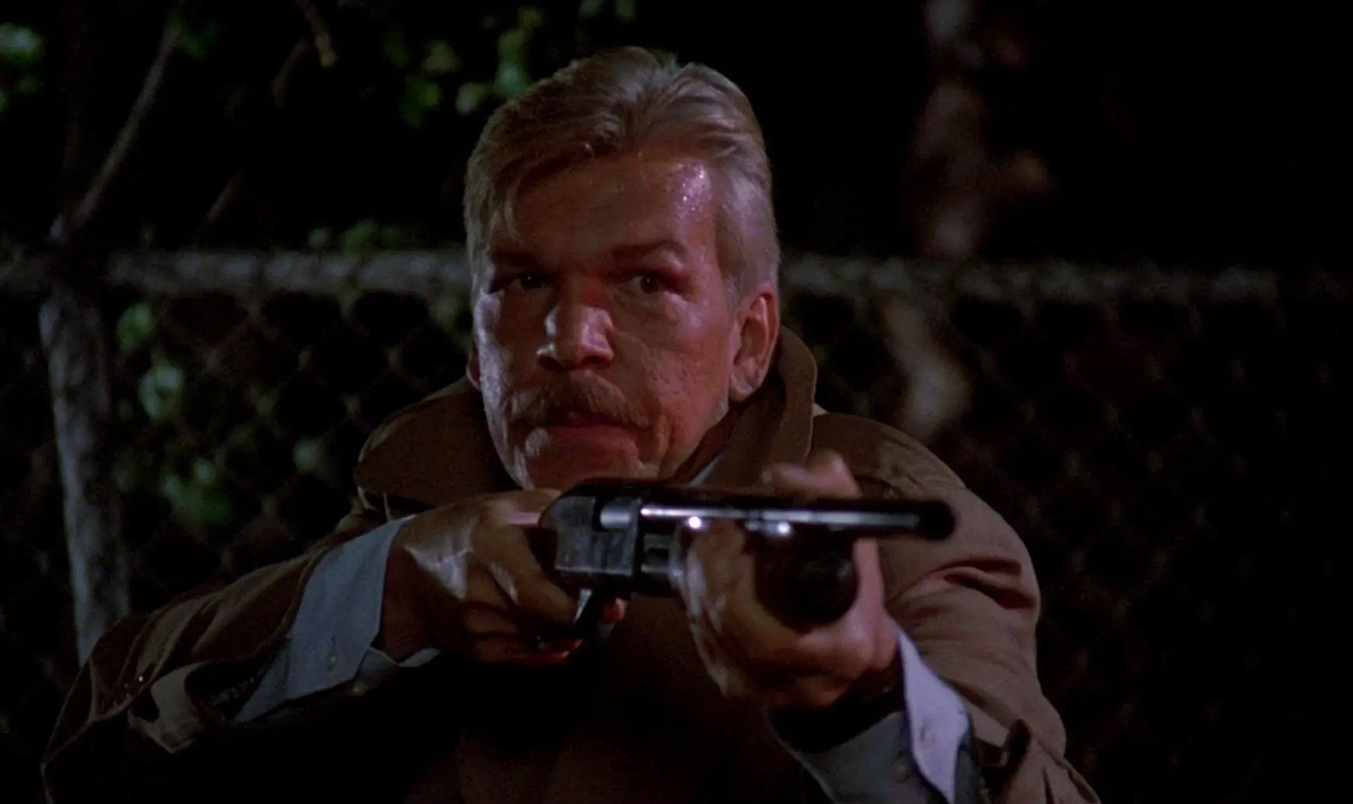 A man with a mustache aims a shotgun while wearing a tan coat at night, with a chain-link fence in the background.