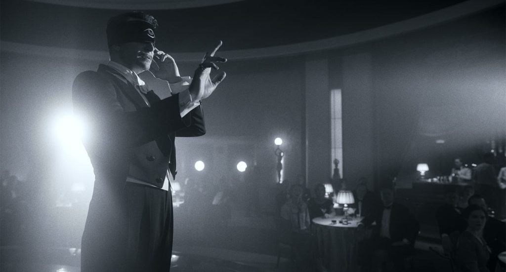 A man in formal attire and a blindfold performs on stage in front of an audience in a dimly lit, elegant venue.