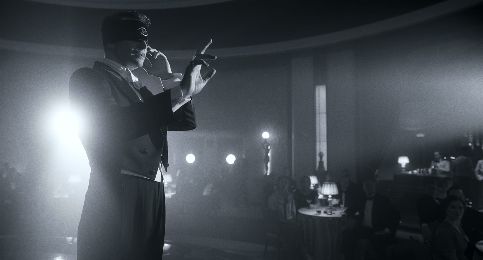 A man in formal attire and a blindfold performs on stage in front of an audience in a dimly lit, elegant venue.