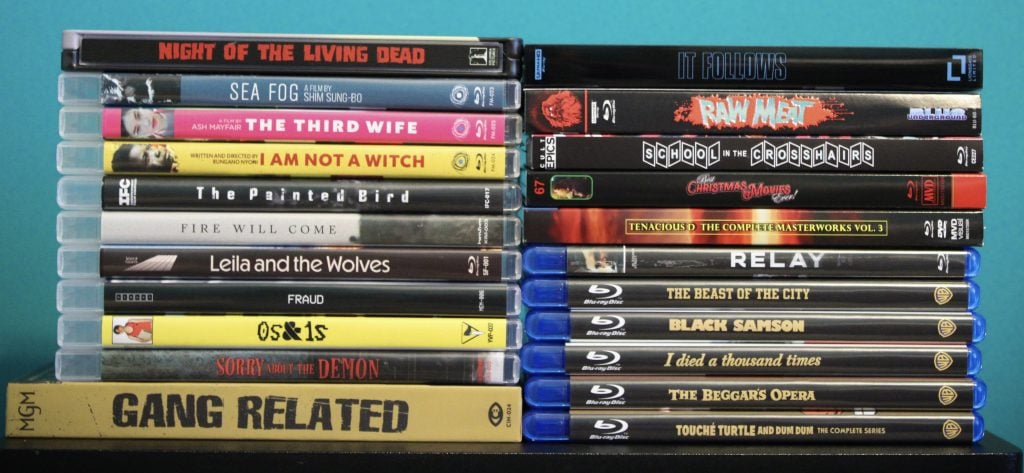 A collection of DVDs and Blu-rays, including titles like "Night of the Living Dead," "It Follows," "Black Samson," and "Gang Related," stacked against a blue background.