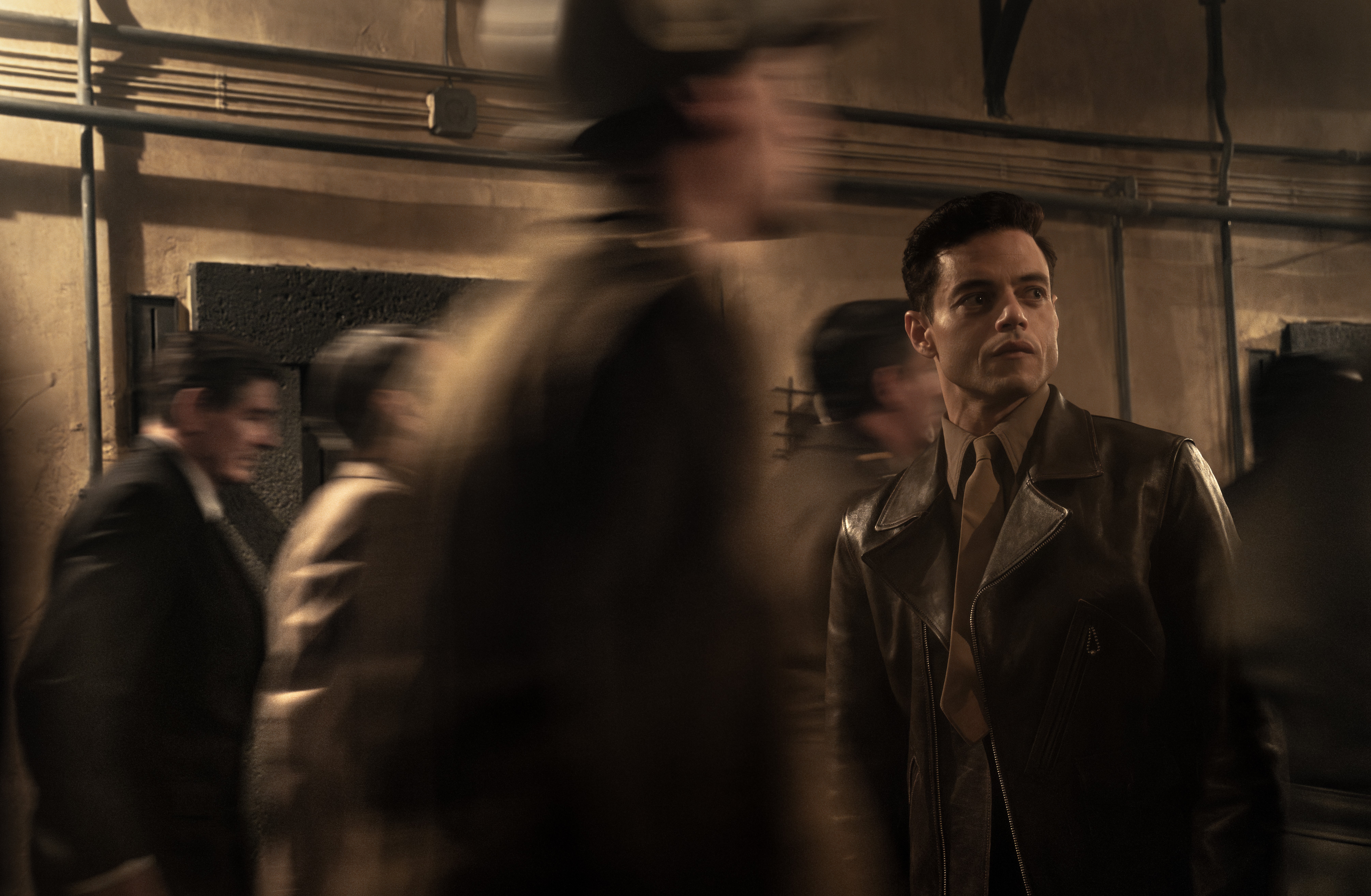A man in a leather jacket stands still while blurred figures walk past him in a dimly lit industrial hallway.