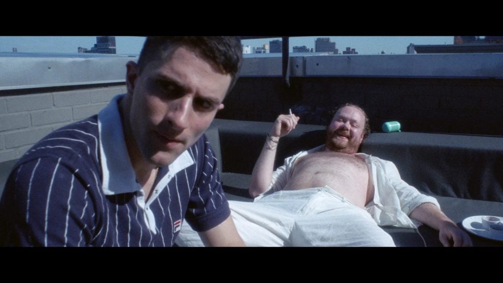 Two men are on a rooftop; one in a striped polo sits close to the camera, while the other reclines shirtless on a couch, smiling and holding a cigarette. Cityscape is visible in the background.