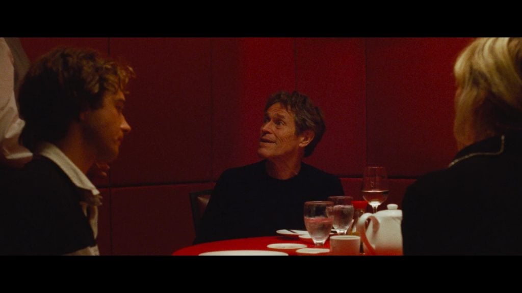 Three people sit at a red table in a dimly lit restaurant, with glasses, plates, and a teapot visible. One man in the center looks up while the others look towards him.