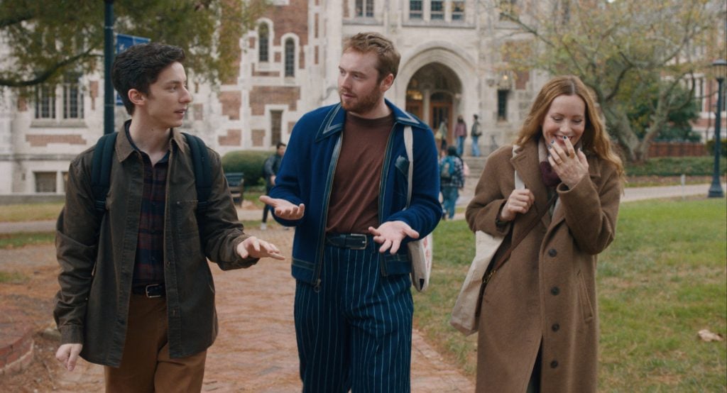 Three young adults walk and talk outside a campus building; one gestures while another laughs, and all carry bags.