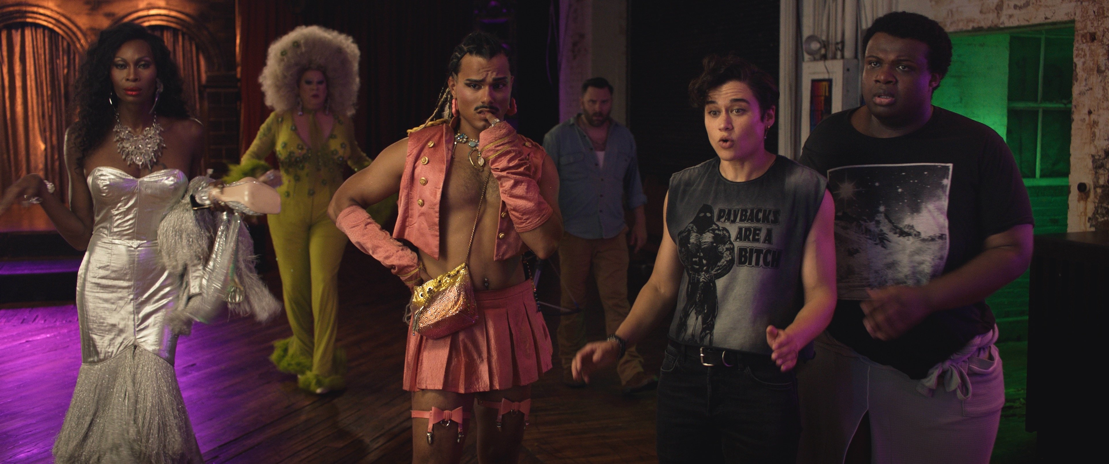 A group of five people stand together in colorful, expressive outfits, with various facial expressions, inside a dimly lit room with wooden floors.