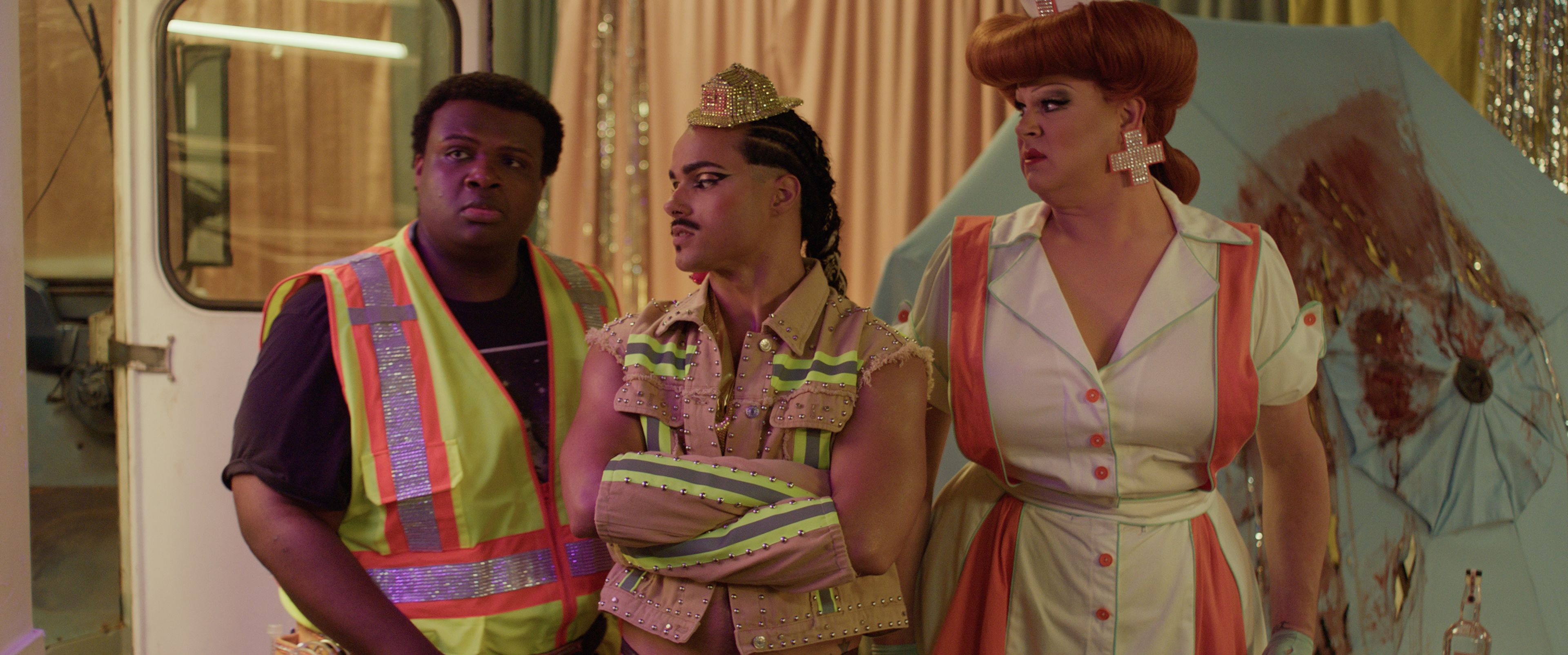 Three people stand together indoors: one wearing a safety vest, one in a decorated outfit with a crown and arm wraps, and one in a retro-style dress with a beehive wig.