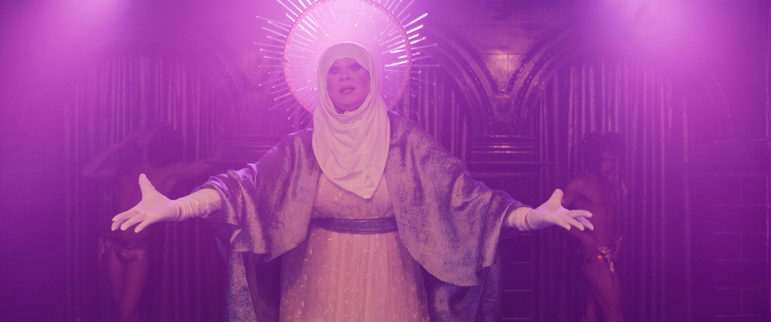 A person wearing a white headdress with a radiant halo, white gloves, and a flowing robe stands with arms open on a dimly lit, purple stage. Two figures are visible in the background.