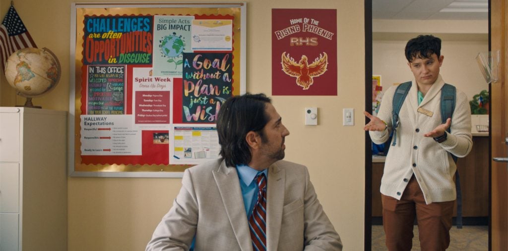 A student with a backpack stands at the door of an office, looking at a man in a suit seated at a desk; colorful posters and a school emblem are on the walls.