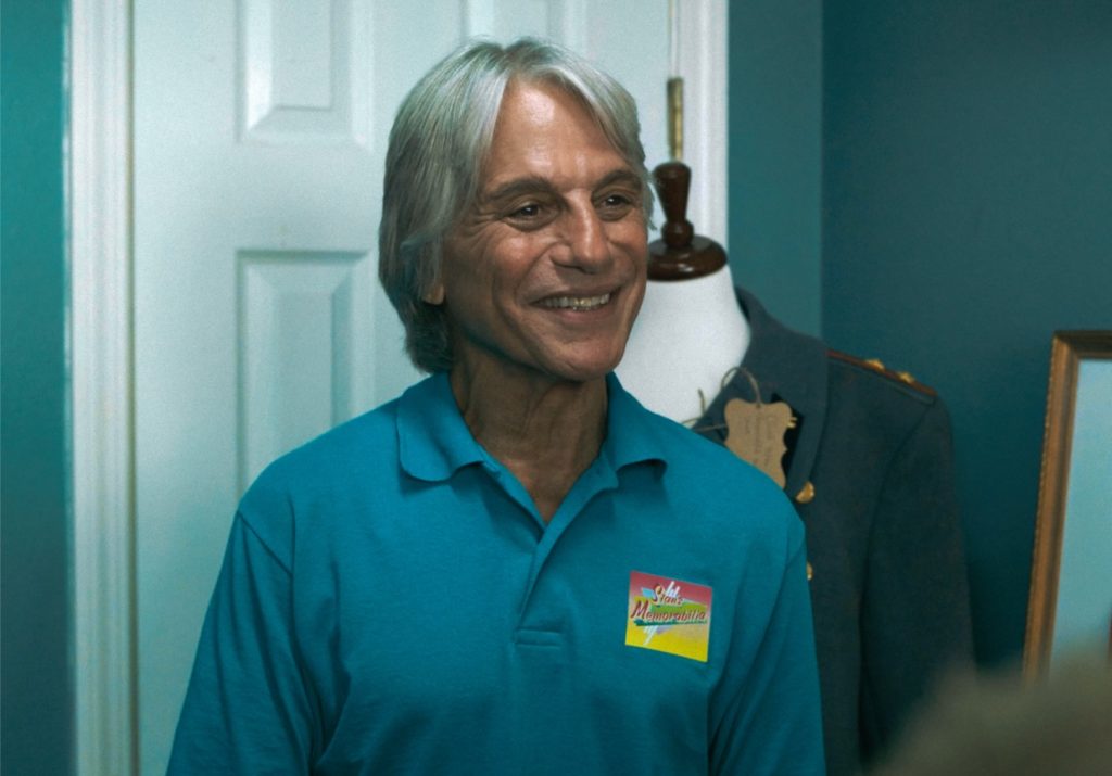 A smiling man with gray hair wears a teal polo shirt with a colorful name tag, standing indoors near a mannequin and a framed picture.