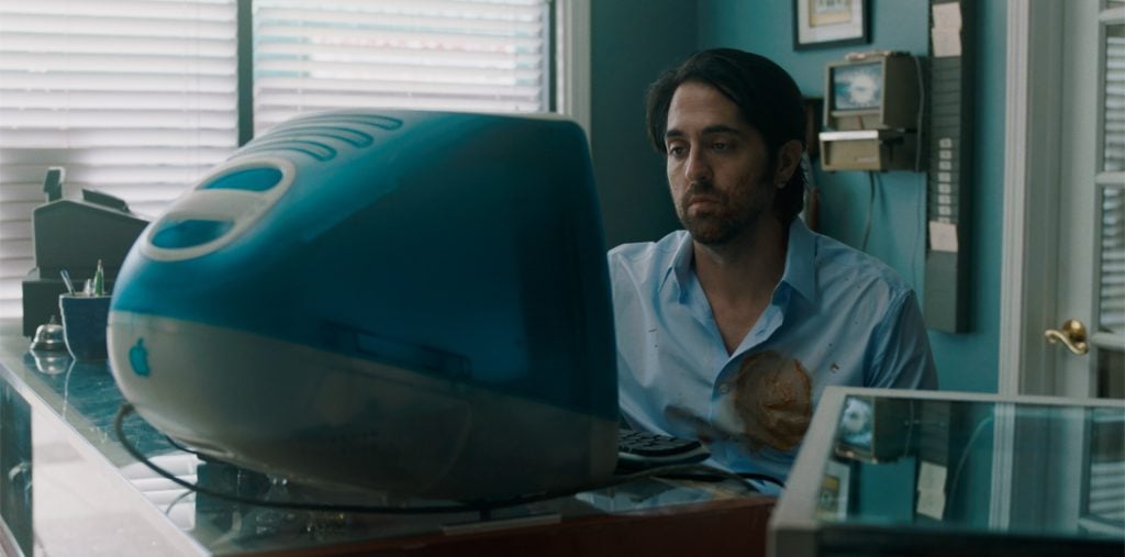 A man sits at a desk in front of a blue iMac computer, wearing a light blue shirt stained with a brownish liquid, in a small office with teal walls.