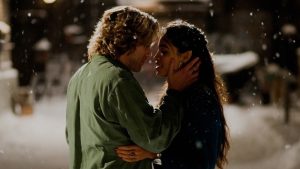 A man and woman stand close together in the falling snow at night, gazing into each other's eyes and about to kiss.
