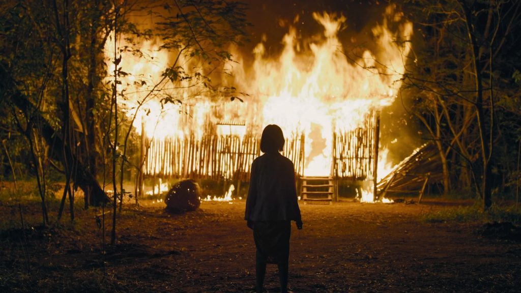 A person stands facing a large wooden structure engulfed in flames at night, with trees visible on either side.