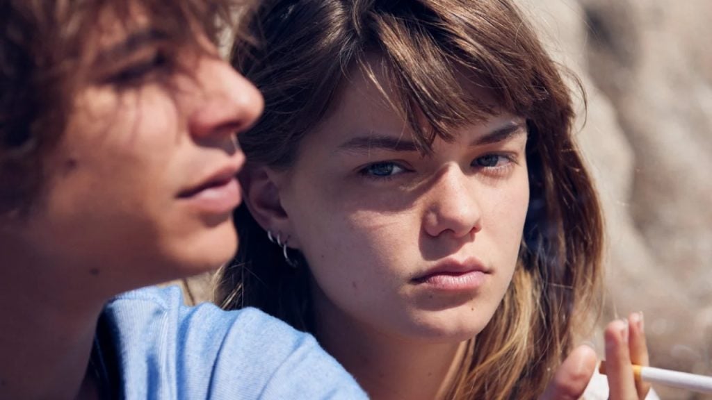 A young woman with light brown hair and blue eyes looks at the camera while holding a cigarette; a young man is out of focus in the foreground.