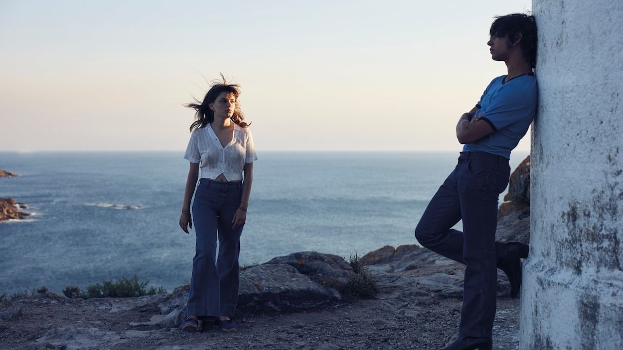 A woman and a man stand near a white wall by the sea at sunset, with the man leaning against the wall and the woman facing him.
