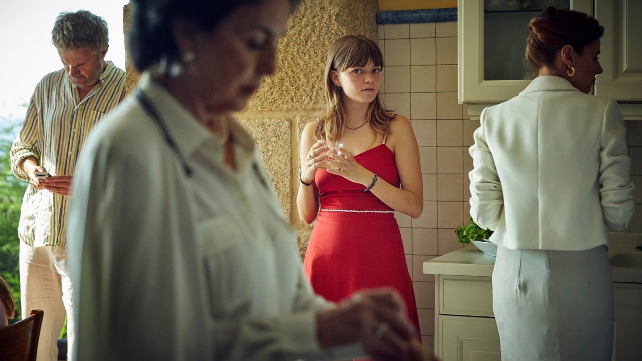 Four people are in a kitchen; a young woman in a red dress stands holding a glass, while others are engaged in separate activities, including using a phone and preparing food.