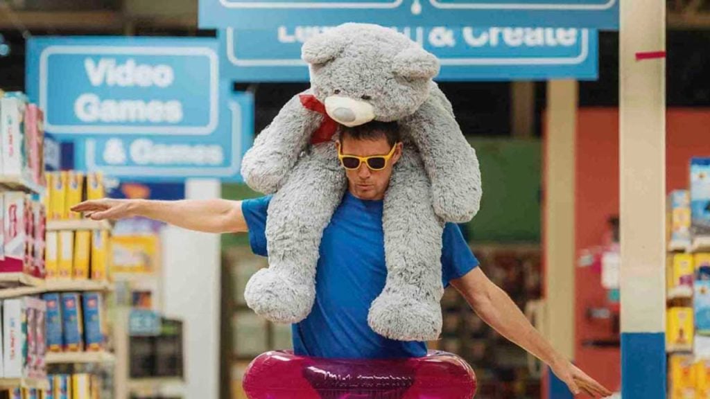 A man wearing sunglasses balances a large gray teddy bear on his shoulders and holds a pink inflatable tube in a store aisle.
