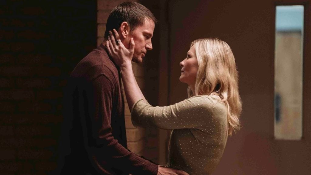 A woman gently holds a man's face with both hands as they look at each other in a dimly lit indoor setting.