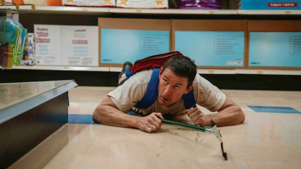 A man wearing a blue vest lies on the floor of a store aisle, holding a grabber tool and looking ahead.