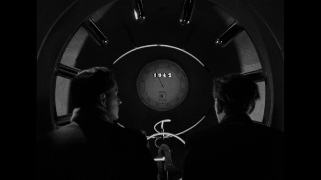 Two people sit facing a circular window showing a clock-like device marked "1942," with metal rods surrounding the window in a dark, shadowed setting.