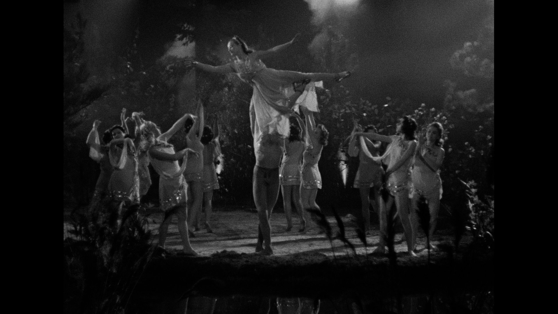 A group of dancers perform on stage; one dancer in a dress is lifted horizontally above the others, who are in matching outfits, under dramatic lighting.