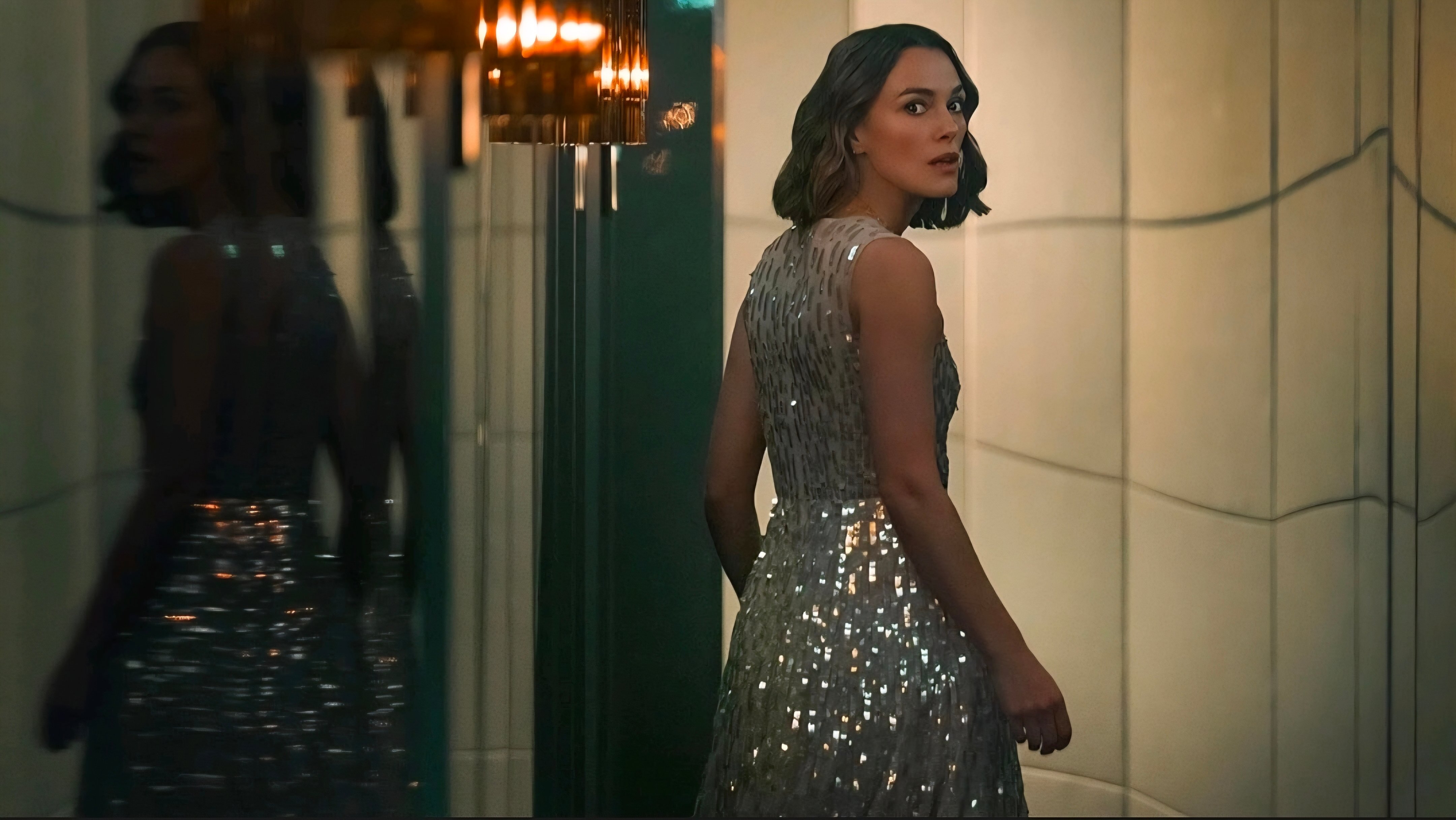 A woman in a sleeveless, silver sequin dress stands indoors, looking over her shoulder at the camera near a reflective wall and a chandelier.