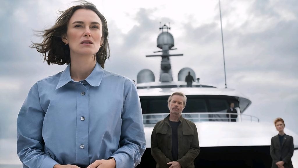 A woman in a blue shirt stands in front of a yacht, with two men in the background on the dock and the yacht's deck, under a cloudy sky.