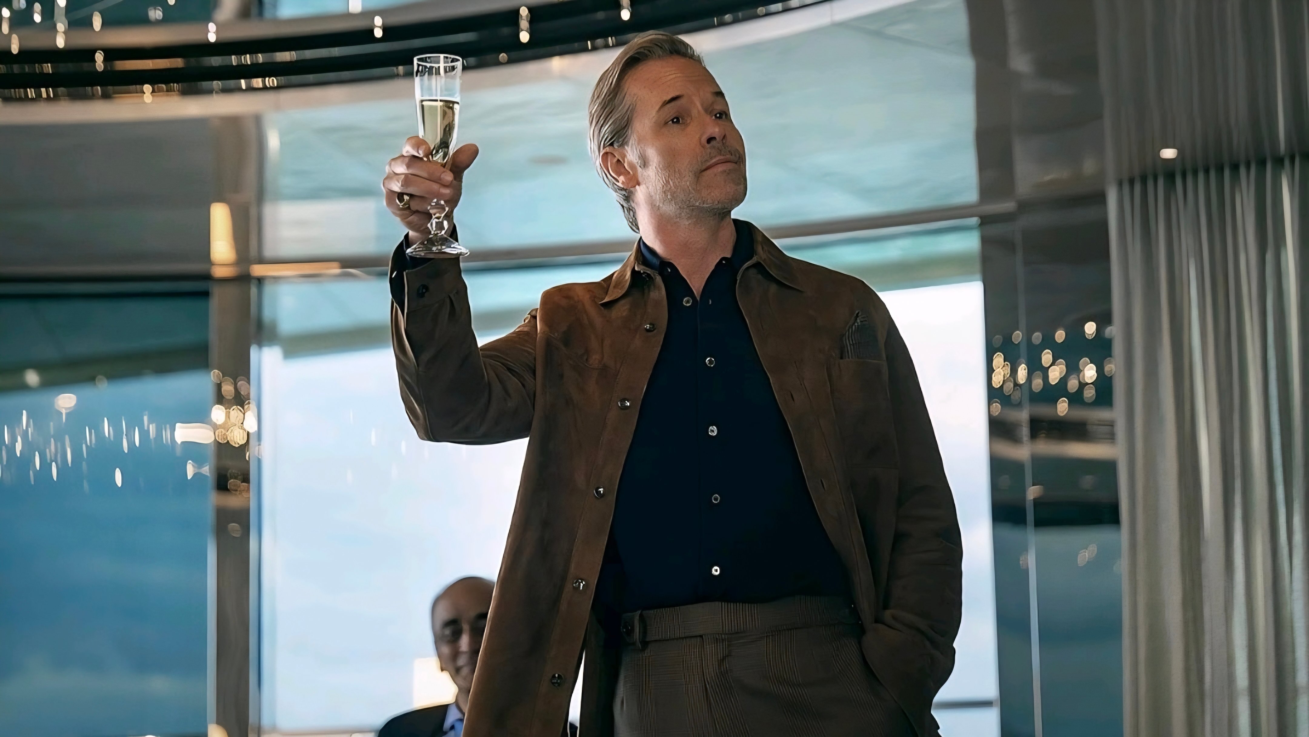 A man in a brown jacket holds up a glass of champagne in a modern, well-lit room with large windows.