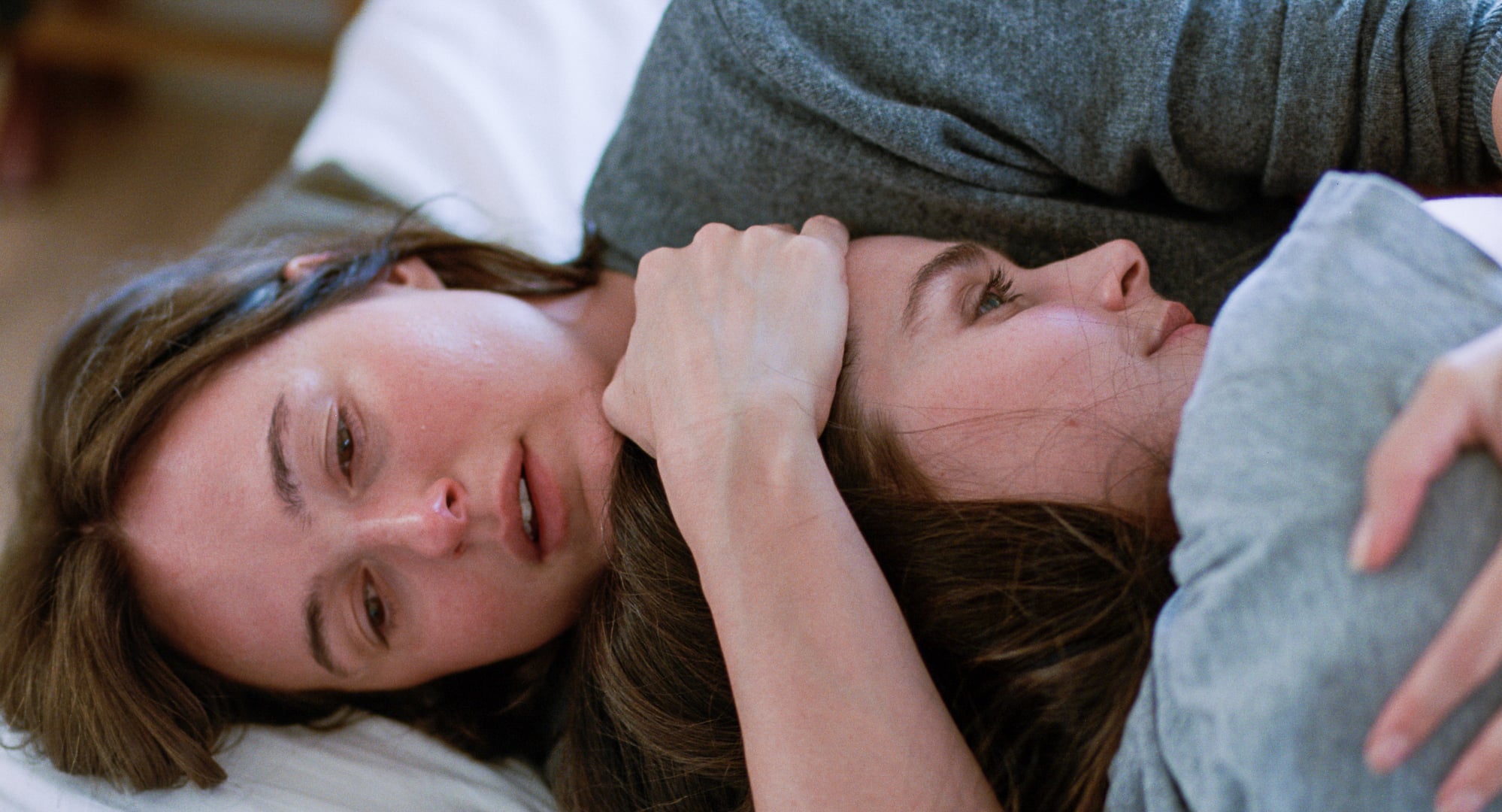 Two women lie close together on a bed, one cradling the other's head. Both have neutral or somber expressions.