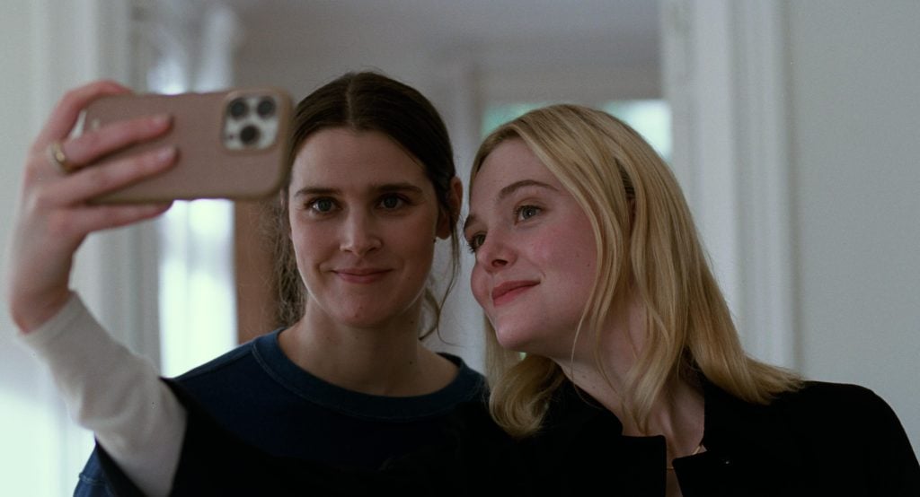 Two young women stand close together indoors, smiling as one of them holds up a smartphone to take a selfie.