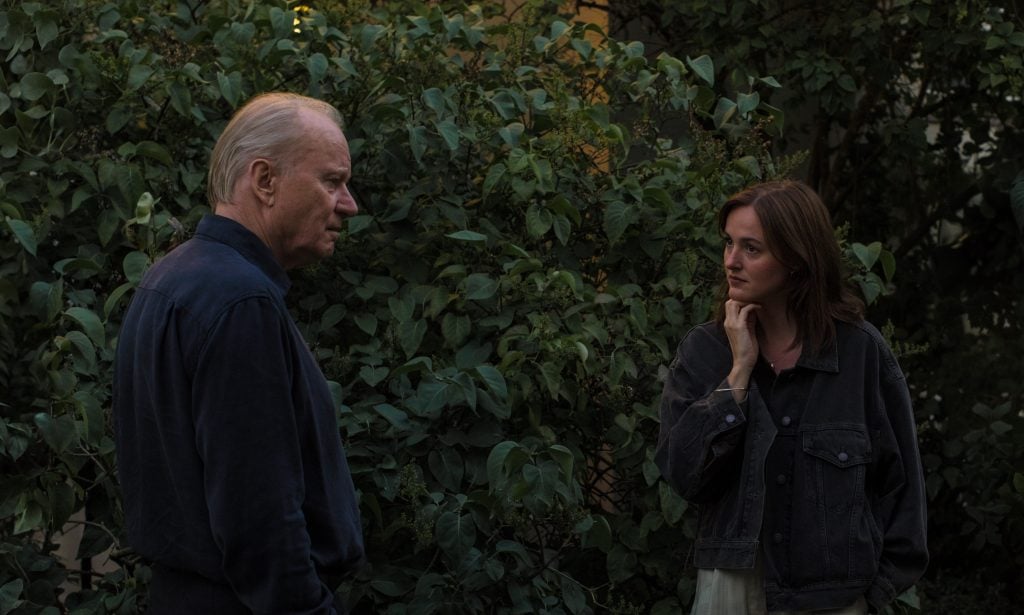 An older man and a young woman stand outdoors in front of dense green bushes, facing each other and engaged in conversation.