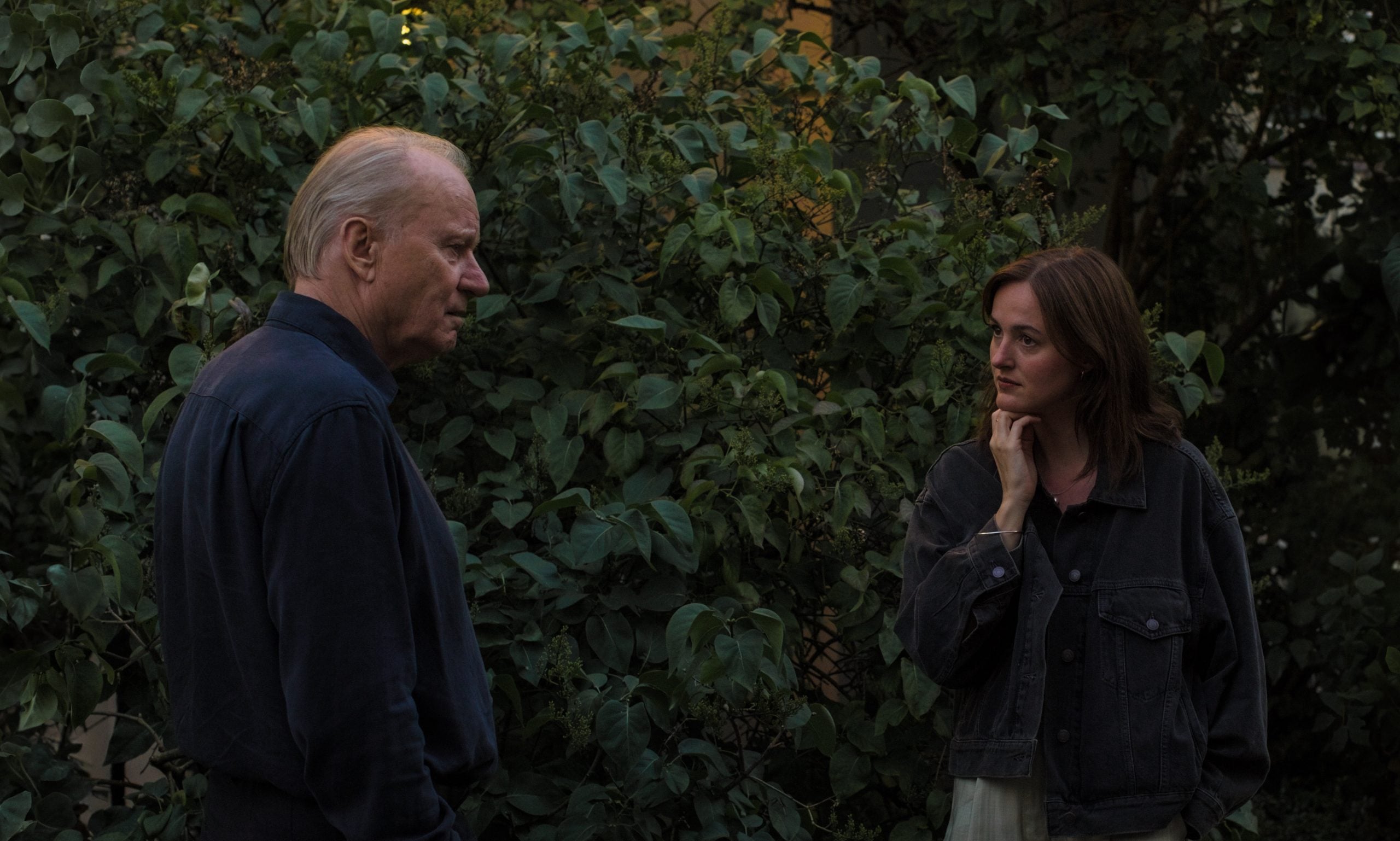 An older man and a young woman stand outdoors in front of dense green bushes, facing each other and engaged in conversation.
