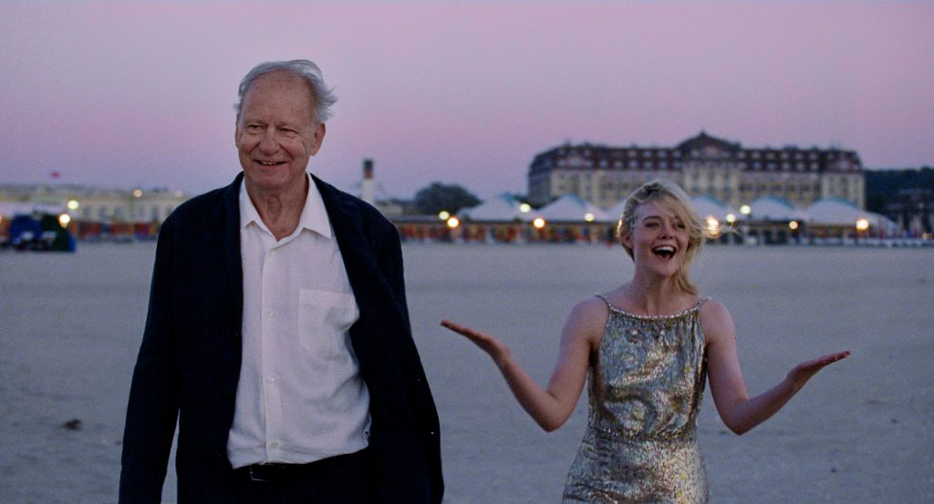 An older man in a dark jacket and a young woman in a silver dress walk on a sandy beach at sunset, with buildings and tents in the background.
