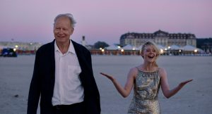 2026 Golden Globe Winner Predictions An older man in a dark jacket and a young woman in a silver dress walk on a sandy beach at sunset, with buildings and tents in the background.