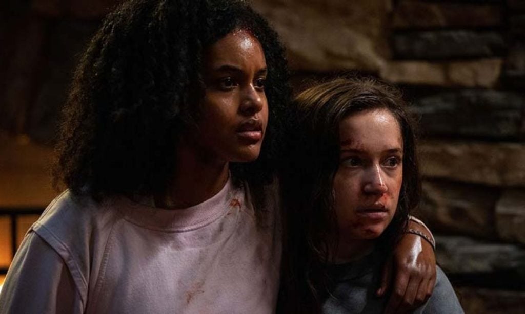 Two young women stand close together, one with her arm around the other. Both have blood on their faces and clothes, appearing tense, with a stone wall in the background.