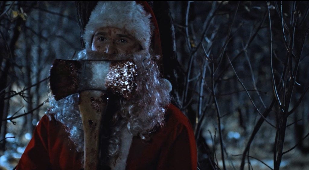 A person dressed as Santa Claus holds a blood-stained axe in a dark, wooded area at night.